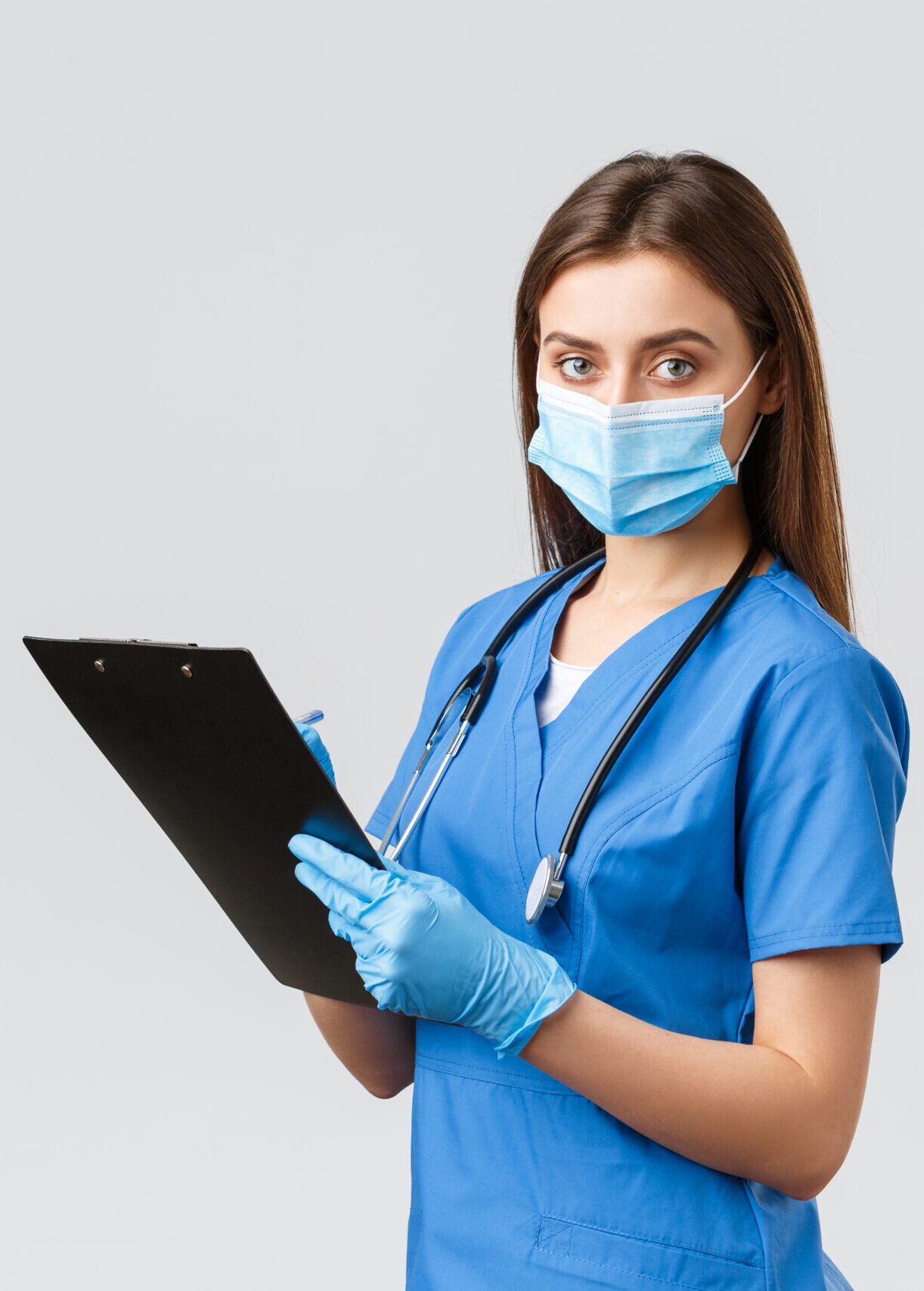 Covid-19, preventing virus, health, healthcare workers and quarantine concept. Professional female nurse or doctor in blue scrubs, medical mask and gloves, writing down patient info using clipboard.
